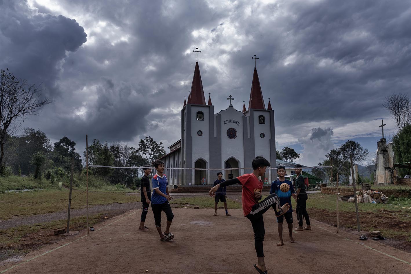 A group of children kick a ball towards a net on a patch of dirt with a church in the background