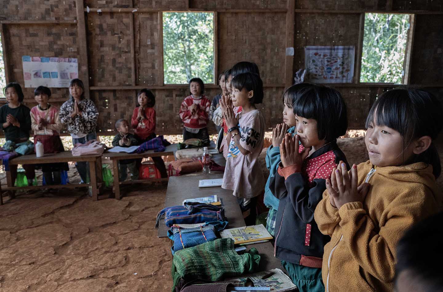 A group of children clasp their hands together while standing behind desks