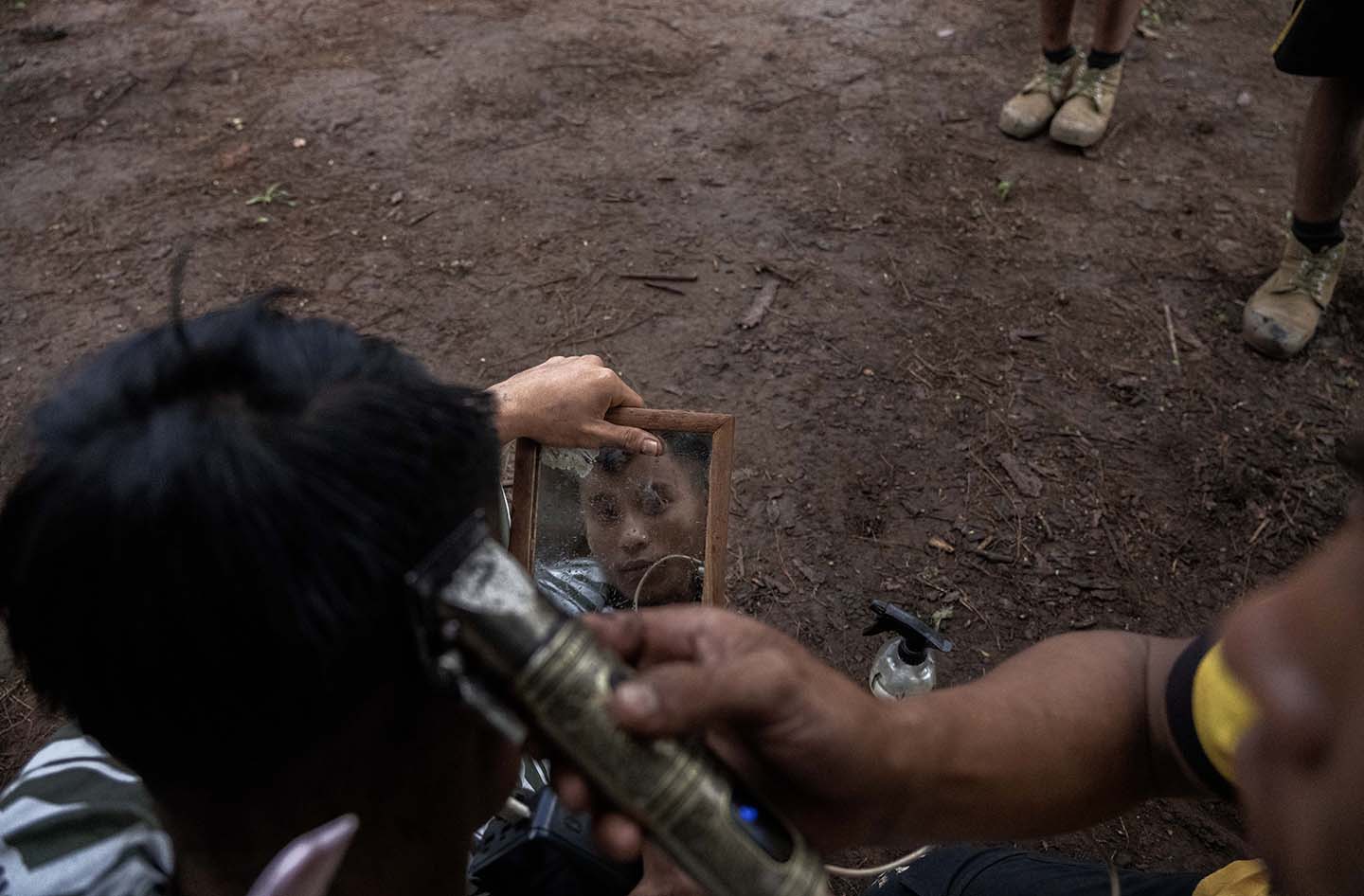 A person stares into a hand mirror as another person shaves their head