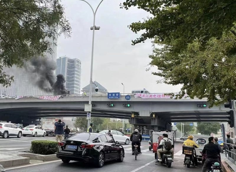 People watch while smoke rises as a banner with a protest message hangs off Sitong Bridge, Beijing, China October 13, 2022 in this image obtained by Reuters.