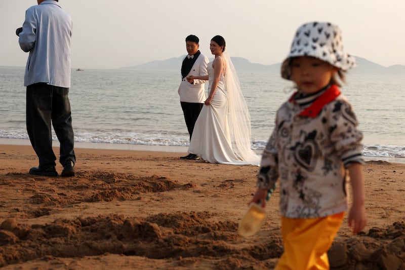 A child plays with sand near a couple taking part in a pre-wedding photo shoot on a beach in Qingdao, China, April 21, 2024.