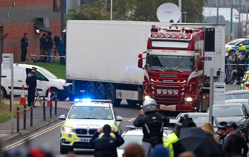 A red semi-truck with a white trailer is escorted by a police vehicle while onlookers snap pictures with smartphones