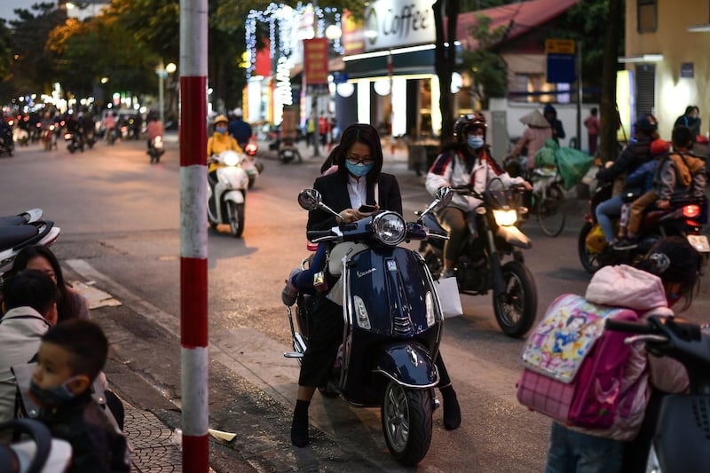 A woman wearing a facemask checks her smartphone while waiting on her scooter along a street in Hanoi, Dec. 1, 2020.