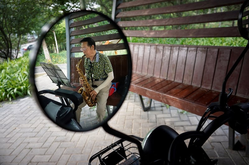 An elderly man is reflected in a rearview mirror of a electric tricycle as he plays a saxophone in a booth for people to rest along a road in Beijing, Aug. 22, 2021. Credit: AFP