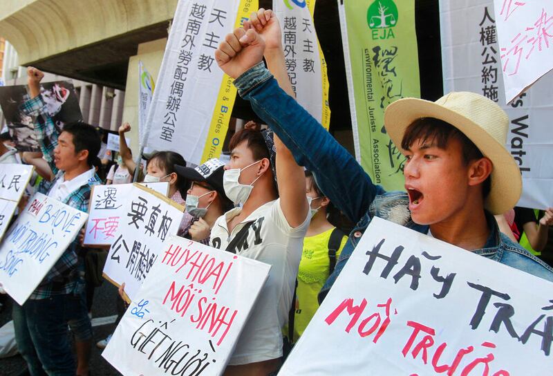 A man wearing a tan fedora raises his right fist and shouts while holding a sign in the right side of the frame. To his right, three other people wear face masks while raising fists and holding signs containing slogans.