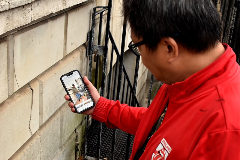 A Vietnamese man in a red zip-up jacket with the Rolling Stones logo on the chest looks down at his smartphone in his right hand, displaying a photo of another Vietnamese man wearing an army green jacket, black baseball cap, and jeans. The man in the photo stands near takeout food that is placed on the ground.