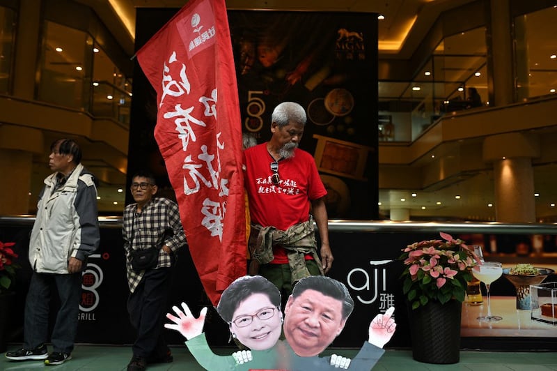 Pro-democracy activist Koo Sze Yiu stands behind placards of Chinese President Xi Jinping and Hong Kong Chief Executive Carrie Lam during a protest before trying to board a ferry to Macau, in Hong Kong on December 18, 2019. The 75-year-old Koo, who has terminal bowel cancer, was concicted on July 12, 2022 of "attempting incitement" of hatred or dissatisfaction of the government under Hong Kong's national security law . Credit: AFP