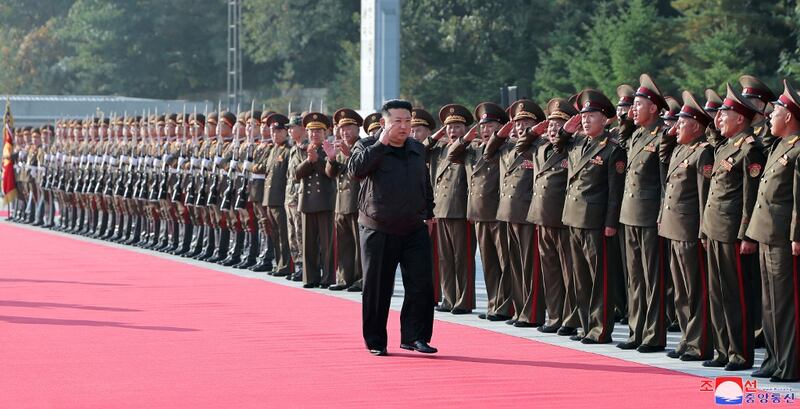 Kim Jong-Un walks on a red carpet in the center of the frame while a long line of soldiers in uniform salute him